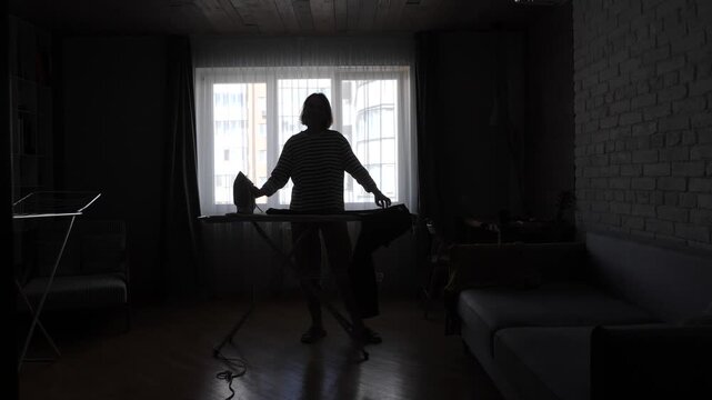 Woman standing in silhouette near a bright window, holding an iron, doing daily laundry tasks on an ironing board inside a dimly lit room, symbolizing routine and domestic labor
