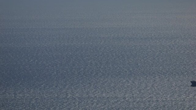 Telephoto view of a cargo ship moving across a shimmering sea surface under soft cloud shadows. Minimalist maritime scene with vast negative space and subtle light variations, ideal for themes of glob