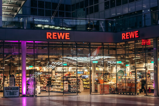 Munich, Germany - April 14, 2025: REWE supermarket at Munich Airport Terminal 2 with illuminated storefront, glass facade and visible interior shelves with food products in evening light