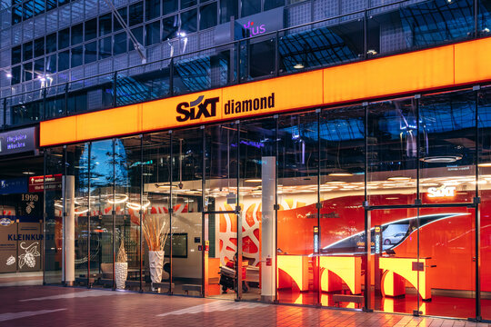 Munich, Germany - April 14, 2025: Sixt car rental office at Munich Airport Terminal 2 with illuminated orange signage, glass facade and customers inside during evening hours