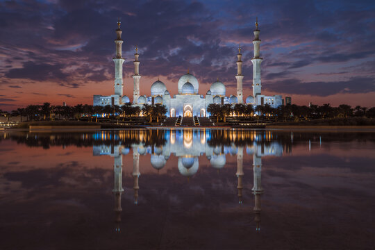 Sheikh Zayed Grand Mosque, illuminated at twilight in Abu Dhabi, UAE, mirrored in a reflecting pool under a dramatic purple and orange sky