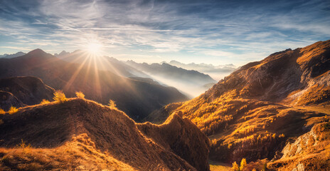 Naklejka premium Mountains in low clouds at beautiful sunset in autumn in Dolomites, Italy. Landscape with alpine mountains, orange grass and trees, hills in fog, blue sky with clouds and sun in fall. Nature