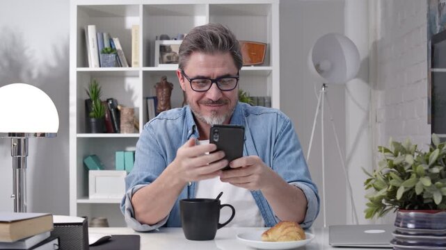 50s man having breakfast checking news on phone drinking morning coffee at home. Confident happy smiling.