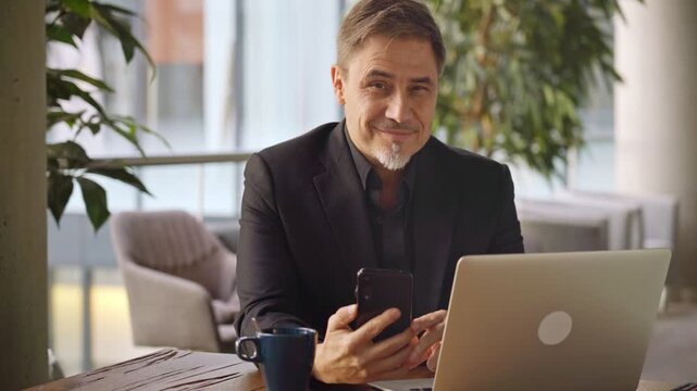 Business portrait - businessman sitting in in office working with laptop computer and phone. Mature age, middle age, mid adult man in 50s with happy confident smile. Copy space.