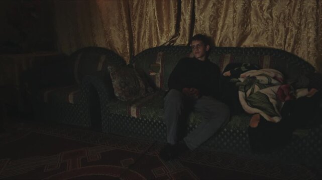 Young man lies down on a couch, covering himself with a warm blanket before falling asleep at night
