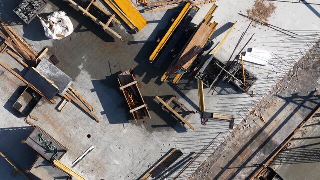 Drone shot of construction materials including formwork panels, wooden planks, and rebar scattered across a concrete slab, showing active site workflow and industrial layout.