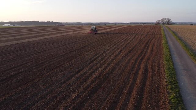 Aerial tracking shot of tractor cultivating soil with low flying drone at sunset