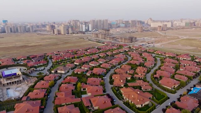 Drone trucks left overhead over gated Ordo cottages with red roofs and green lawns, revealing curved streets, roundabouts, private pools and distant Bishkek skyline on the hazy horizon amid suburban
