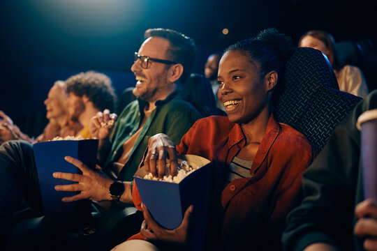 Happy black woman eating popcorn while watching movie at cinema.
