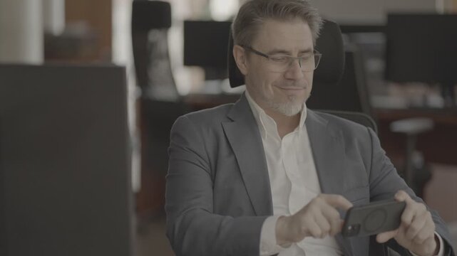 Business portrait - confident businessman sitting at desk working in office, using phone. Happy mid adult man in shirt and jacket, smiling. Video in LOG format - S-LOG3 Cine.