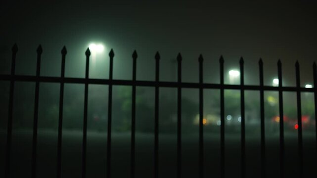 Metal fence in the foreground at night with street lights and urban lights blurred by dense fog in the background, creating a dark and atmospheric city scene