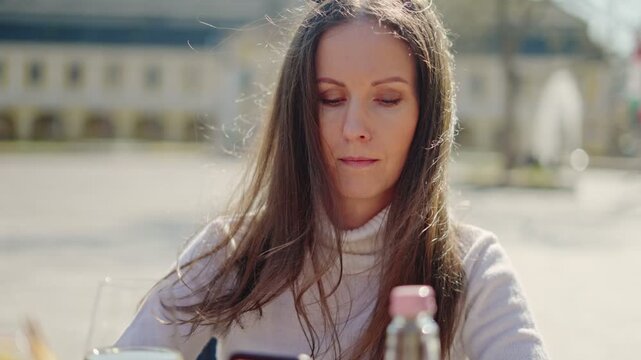 Woman sitting outdoor on terrace in restaurant or cafe, using phone, posting on social media. Wind blown hair, spring or autumn, sunny day.