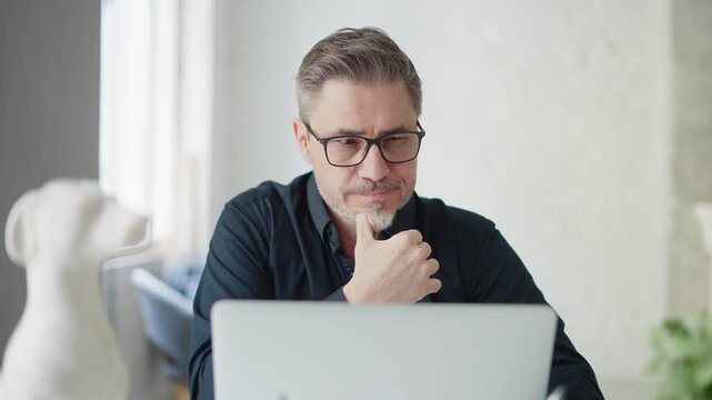 Business portrait - confident businessman sitting at desk in office working with laptop computer. Happy mid adult man in shirt and jacket, smiling.