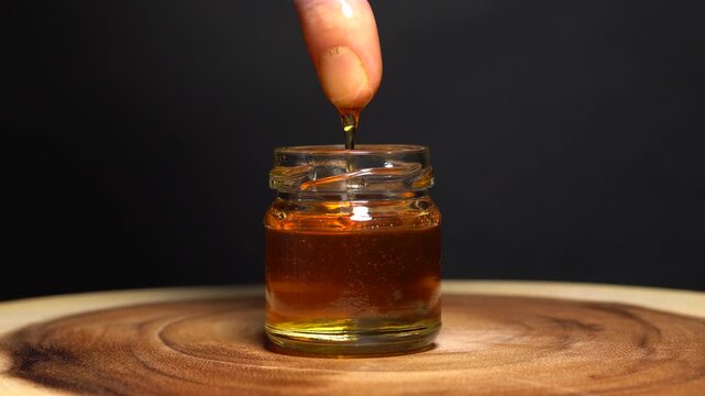 Dipping finger into a jar of natural honey, macro close up selective focus. Man dips his finger into bee honey in the can on the wooden table or tree trunk log. Beekeeping and product quality testing