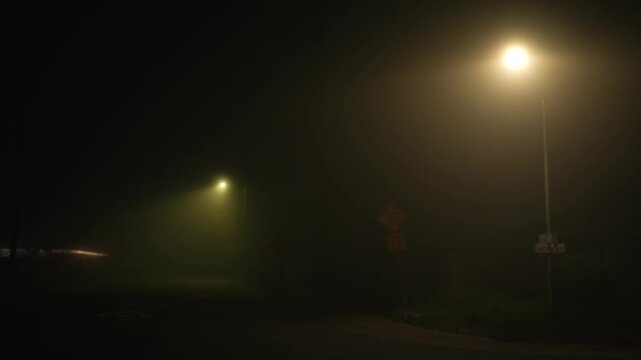 Urban street at night covered in dense fog, with street lights softly diffusing through the mist and a barely visible road sign in the background, creating an atmosphere of silence and uncertainty