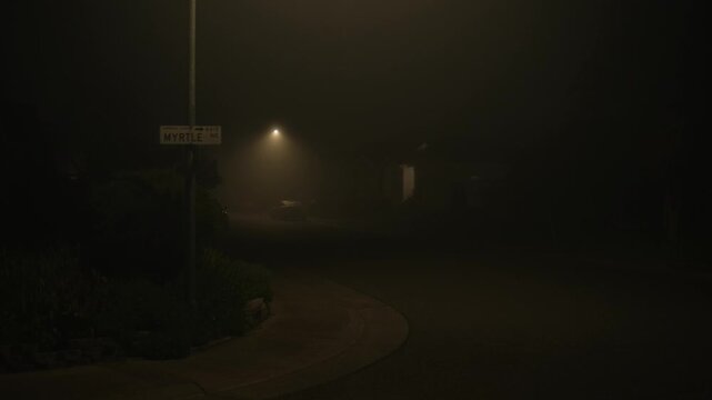 Residential street at night covered in dense fog, with a soft glowing street light and a barely readable street name sign, creating a sense of silence, isolation, and mysterious atmosphere