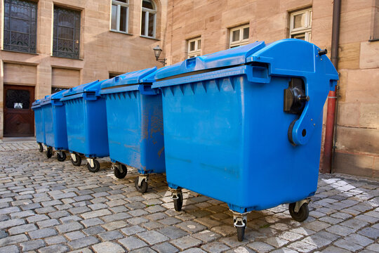 Row of large blue plastic garbage bins on wheels standing on a cobblestone street. Concept of waste management, recycling, and urban cleanliness.