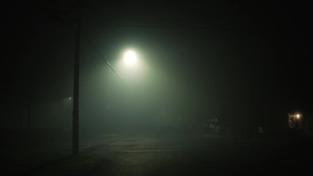 Nighttime intersection covered in dense fog, where the bright glow of a street light diffuses through the mist, highlighting overhead power lines and an empty road, creating an atmosphere of silence
