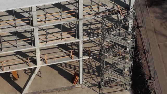 Drone shot of a multi-level concrete building frame with scaffolding, stairs, and organized construction materials on-site, highlighting structural development and engineering progress.