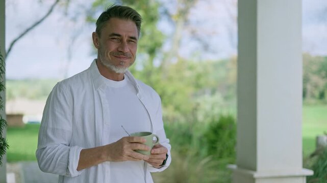Relaxed businessman standing on porch at home, holding mug, looking away. Mature man in white shirt drinking morning coffee on terrace. Male in 50s. Tranquility of country life, stressless lifestyle.