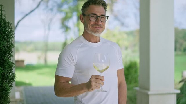 Mature man drinking white wine on porch of country house. Happy middle aged male in white outfit standing outside on ranch terrace tasting from glass, smiling. Tranquility and countryside lifestyle.