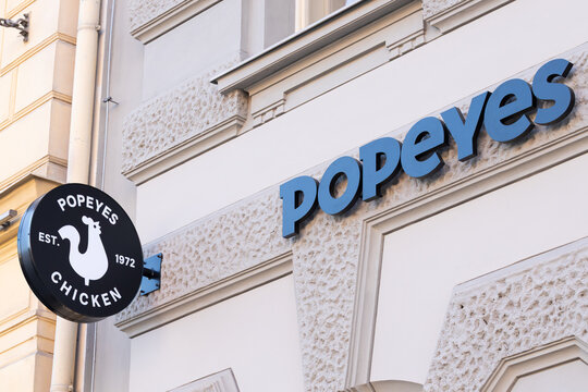 Restaurant exterior featuring a prominent black sign with the name Popeyes and a round logo displaying a chicken silhouette, established in 1972, on a decorative building facade