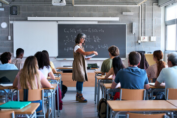 Adult female teacher stands by the blackboard explaining a science formula to a diverse group of students in a modern classroom during a lesson. © CarlosBarquero