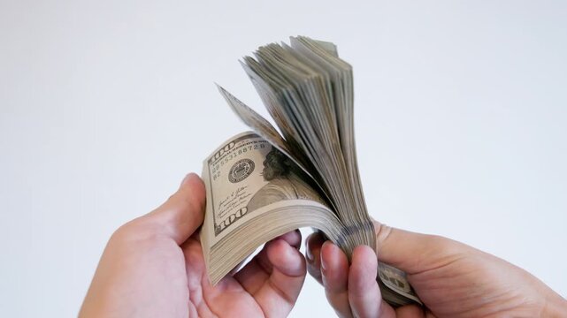 Two hands methodically count a large stack of one hundred dollar bills against a plain white background. Neutral lighting enhances the scene of finance and wealth.