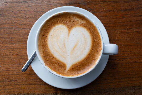 Heart-shaped latte art on a creamy cappuccino in a white ceramic cup, served on a wooden table. Top view of a cozy morning coffee break with a romantic foam design.
