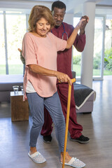 Naklejka premium Senior woman practicing walking with cane, African American caregiver supporting in scrubs at home