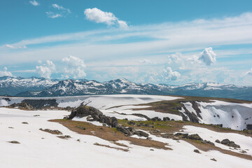 Scenic alpine landscape with sunlit snowy field on stony hill with view to big mountain range with forest and snow far away under cloudy sky. Stone outcrops among snows in sunlight in high mountains. © Daniil