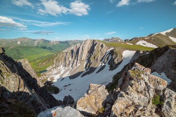Aerial dizzying view to rocky cliff between sharp rocks under clouds in blue sky. Vertigo scenery with sheer crags above small lake in green dale and mountain top far away in cloudy changeable weather © Daniil