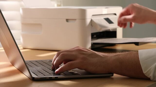 Man using printer while working on laptop at wooden desk indoors, closeup