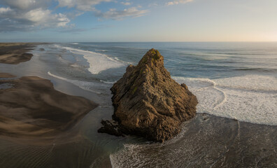 Obraz premium Aerial view of the iconic Rock at Karekare Beach, New Zealand. Waves crash around the rock formation, showcasing the rugged beauty of the coastline in Auckland.