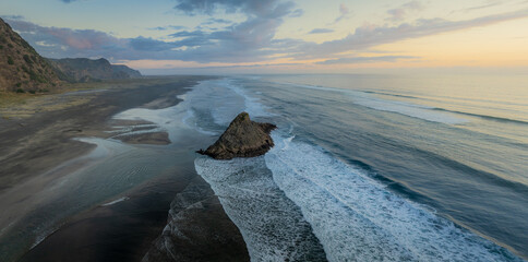 Obraz premium Aerial view of the black sands of Karekare Beach, New Zealand, with a Rock standing tall against the crashing waves. A popular spot for surfers and nature lovers.