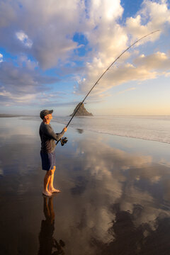 A man fishes at sunset on the beach in Karekare, New Zealand. He is surfcasting with a long rod, hoping to catch a fish for dinner. The sky reflects on the wet sand.