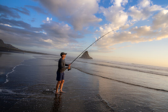 Auckland, New Zealand: A man fishes at sunset on the beach at Karekare. He is surfcasting with a long rod, hoping to catch fish for dinner, enjoying the outdoors.