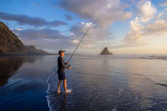Auckland, New Zealand: A man fishes at sunset on the beach at Karekare. He is surfcasting with a long rod and reel, hoping to catch fish for dinner.