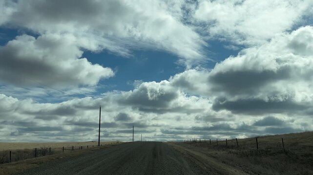 POV driving along rural gravel road beneath dramatic cloud break with blue sky opening and power lines crossing open prairie landscape