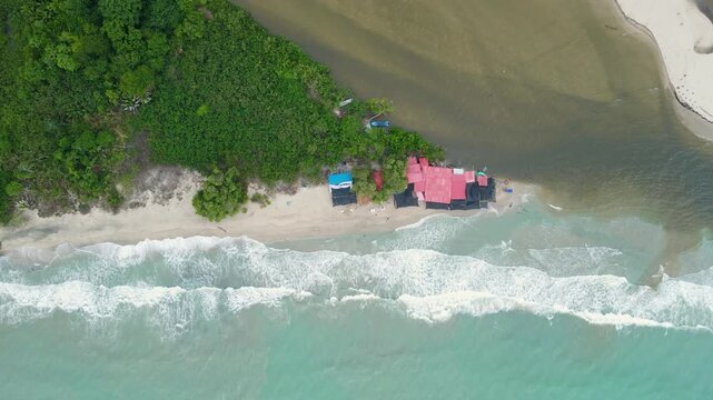 Drone view of red-roof beach huts and a blue boat as turquoise surf and brown river water mix along a narrow sandy shoreline in Palomino.
