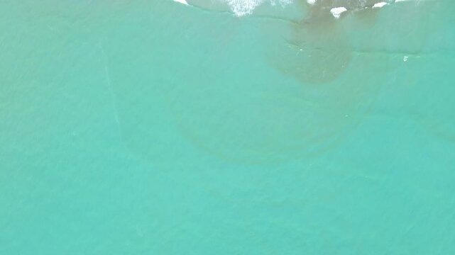 Top-down drone view of turquoise sea and breaking waves mixing brown sediment at a sandbar, with red-roof houses and green vegetation along the shoreline.