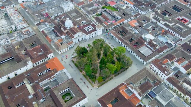 Drone footage over a tree-filled central square in Popay&aacute;n, showing intersecting walkways, a white church dome and tiled rooftops as people move through the plaza.