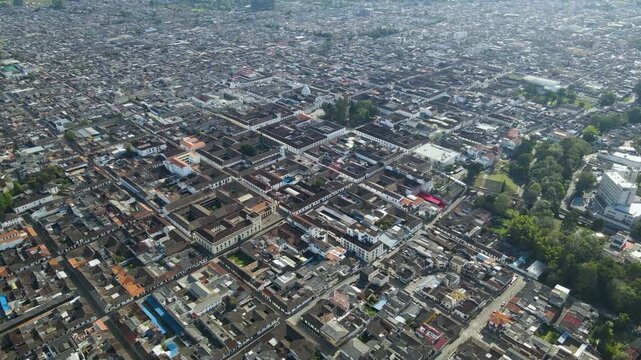 Drone flyover of Popay&aacute;n showing white colonial buildings, red tile roofs and a tree-lined park within the city grid under bright daylight.