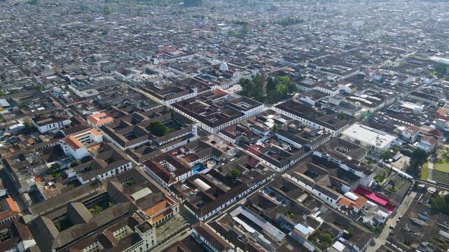 Aerial drone sequence over Popay&aacute;n showing white colonial buildings, clay tile rooftops and a central church dome, with grid streets and tree-lined blocks under hazy daylight.