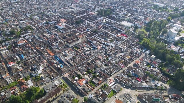 Aerial shot over Popayan historic center showing grid streets, tiled rooftops and a tree-lined park bathed in daylight as the drone slowly sweeps across the city.