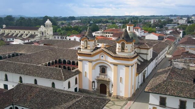 Drone approaches a white colonial church with yellow trim and green dome, revealing tiled roofs, a cloister courtyard and a quiet cobblestone plaza under a cloudy daytime sky.