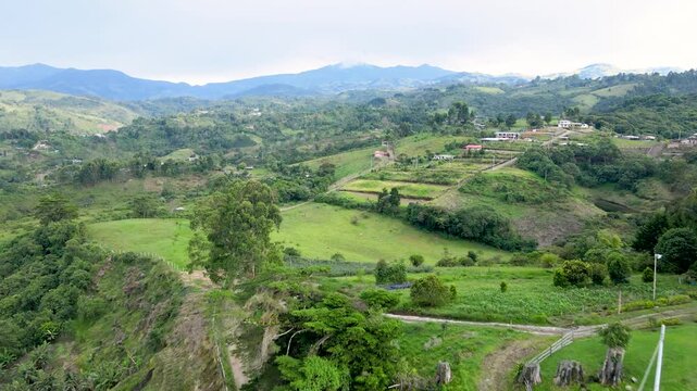 Drone pans over green hills and terraced fields near Popay&aacute;n, revealing a winding dirt road and scattered red-roof houses beneath a cloudy mountain skyline.