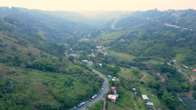 Drone footage of a winding Pan-American road through a green valley near Popayan, with scattered houses, passing trucks and a small river under hazy daylight.