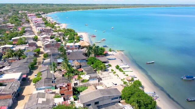 Drone footage over Rincon del Mar shows turquoise Atlantic water and a curved sandy beach with moored fishing boats, palm trees and thatched roofs along the village.