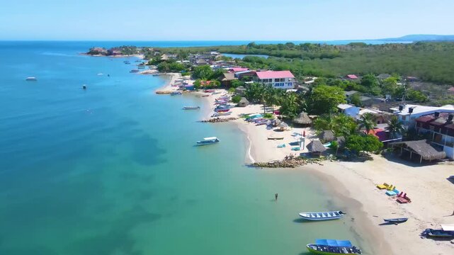 Drone flight over Rincon del Mar reveals turquoise Atlantic water, sandy beach with small boats and red-roof houses beside dense green mangroves under a clear blue sky.